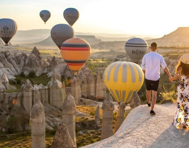 Sunrise hot air balloons floating over the fairy chimneys of Love Valley in Cappadocia, with two people standing in the foreground enjoying the panoramic view